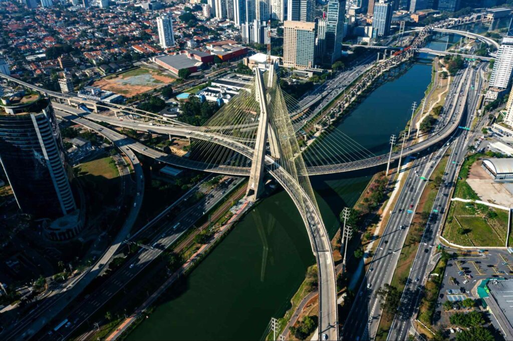 Imagem aérea das pontes e viadutos que cruzam o Rio Tietê, em São Paulo. De ambos os lados os prédios e casas da cidade, além de pistas com carros trafegando.