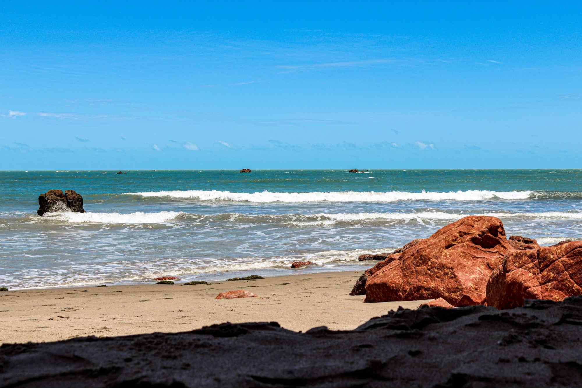 Grandes pedras na areia da praia de Icapuí, em frente ao mar, com céu azul e limpo. O mar se estende até o horizonte, e mais algumas pedras podem ser vistas com as pontas para fora do mar.