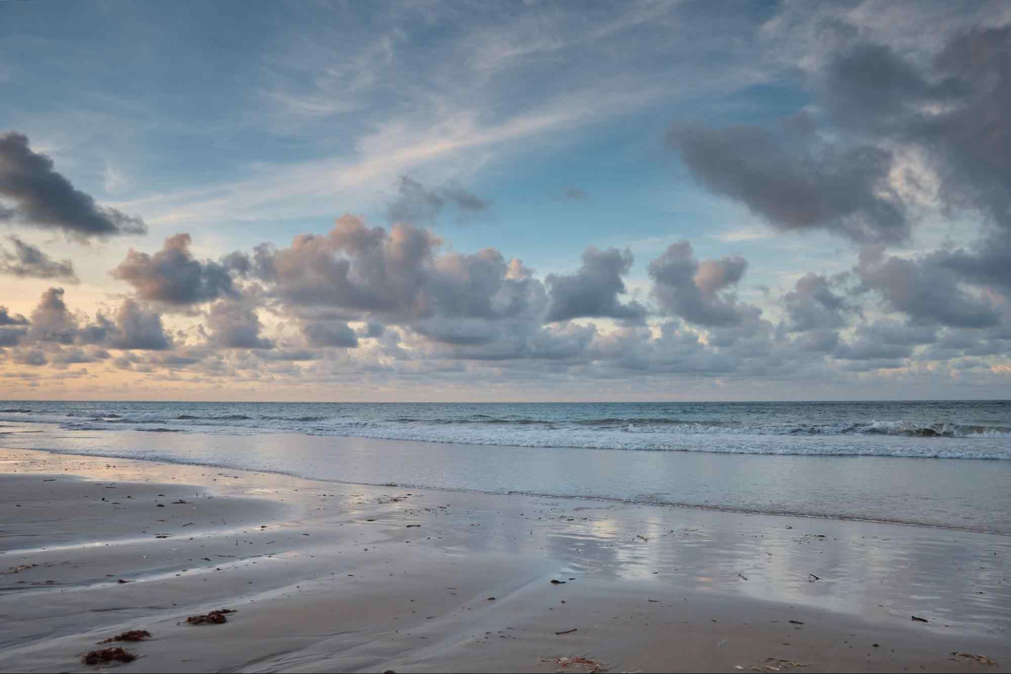 Praia em Paracuru. A foto mostra o mar, que é claro e sem ondas, e um trecho de areia molhada. O céu está dividido entre tonalidades de azul e dourado, por causa do pôr do sol.