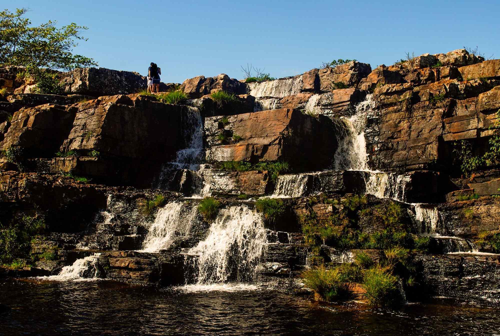 O enorme paredão de pedra por onde despenca a Cachoeira Grande, localizada na Serra do Cipó MG. No alto do cachoeira, um casal posa para uma foto.