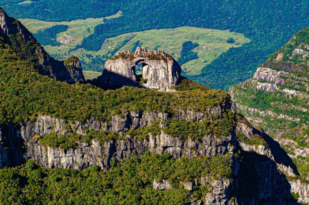 Formação rochosa Pedra furada, com uma abertura circular no meio, no topo de um monte. A montanha de pedra cinzenta tem trechos cobertos por árvores e, ao fundo, é possível ver uma mata densa.