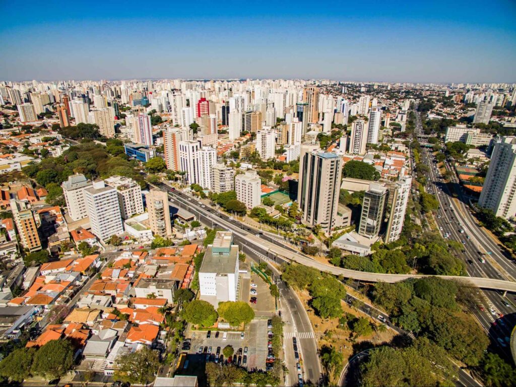 Vista panorâmica da Vila Mariana, em São Paulo. A paisagem urbana é composta por duas grandes avenidas, muitos prédios altos e ruas ricamente arborizadas. Ao fundo, aparece o céu sem nuvens.