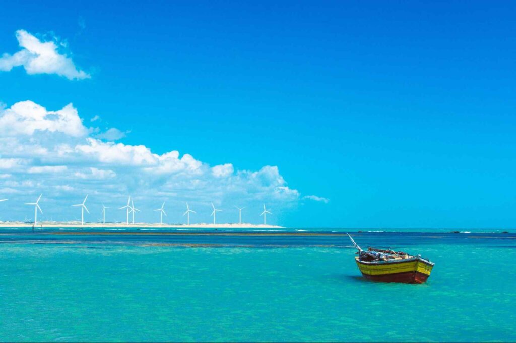 Mar da Praia de Icaraizinho de Amontada. A água é de uma tonalidade azul vibrante, similar à cor do céu limpo, e se estende até o horizonte. Em primeiro plano, aparece um pequeno barco amarelo que flutua na água.