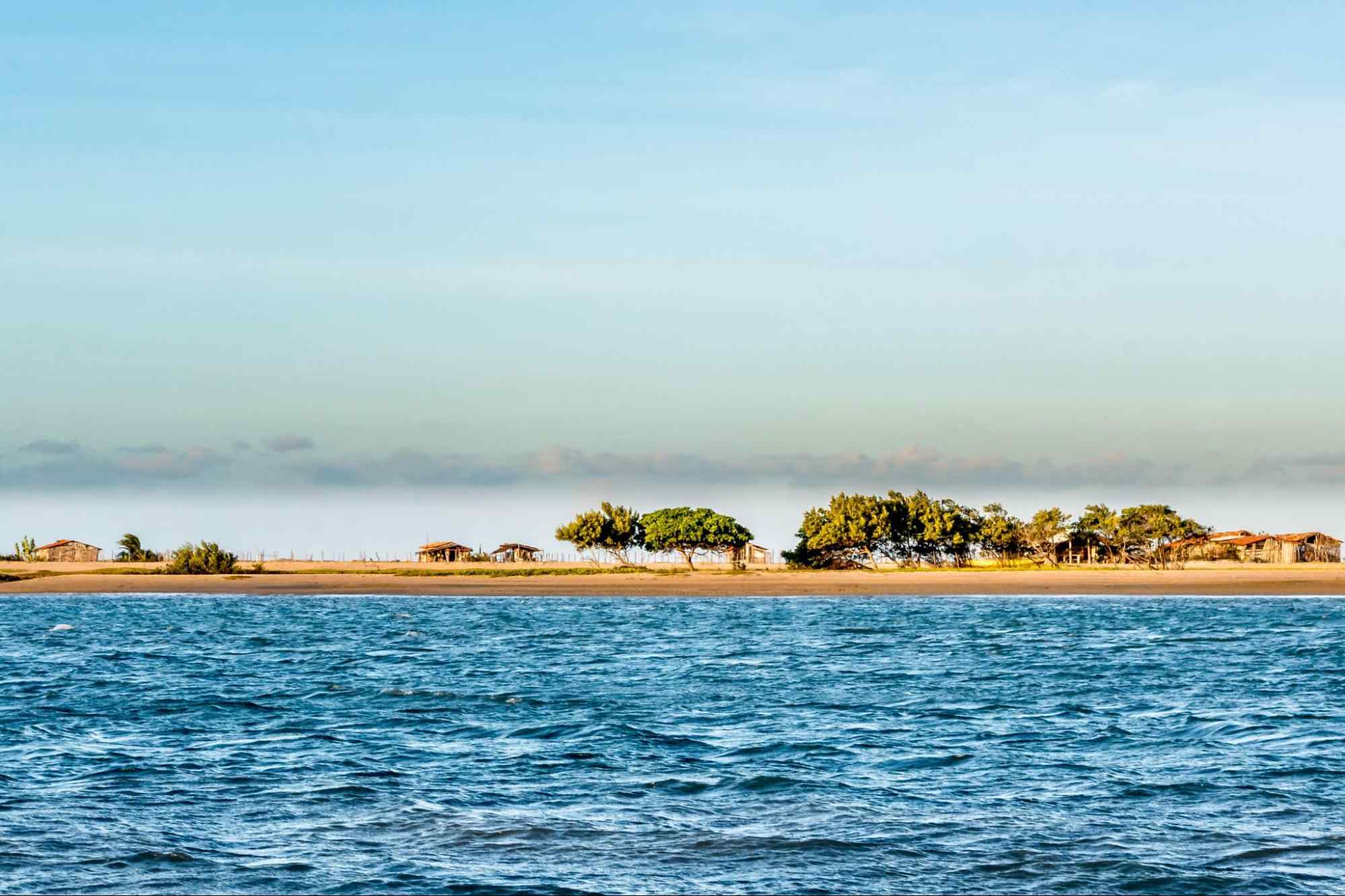 Praia de Guajiru em Trairi CE, vista do ponto de vista do mar. Ao fundo está a faixa de areia com algumas árvores e pequenas casas, enquanto o mar é de um azul intenso.