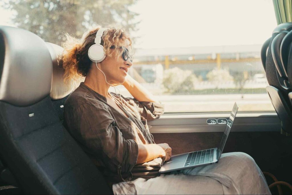 Mulher jovem sentada em uma poltrona de ônibus de viagem. Ela usa fones de ouvido e óculos escuros e tem um laptop aberto apoiado sobre uma das pernas.