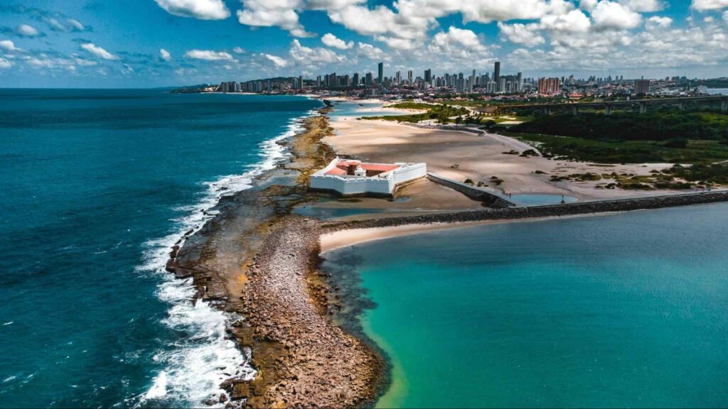 Imagem aérea do encontro entre o Rio Potengi e o mar, separados por uma faixa de areia rochosa, com o Forte dos Reis Magos entre as águas. Mar azul com ondas brancas, Rio esverdeado em dia de céu azul com várias nuvens brancas.