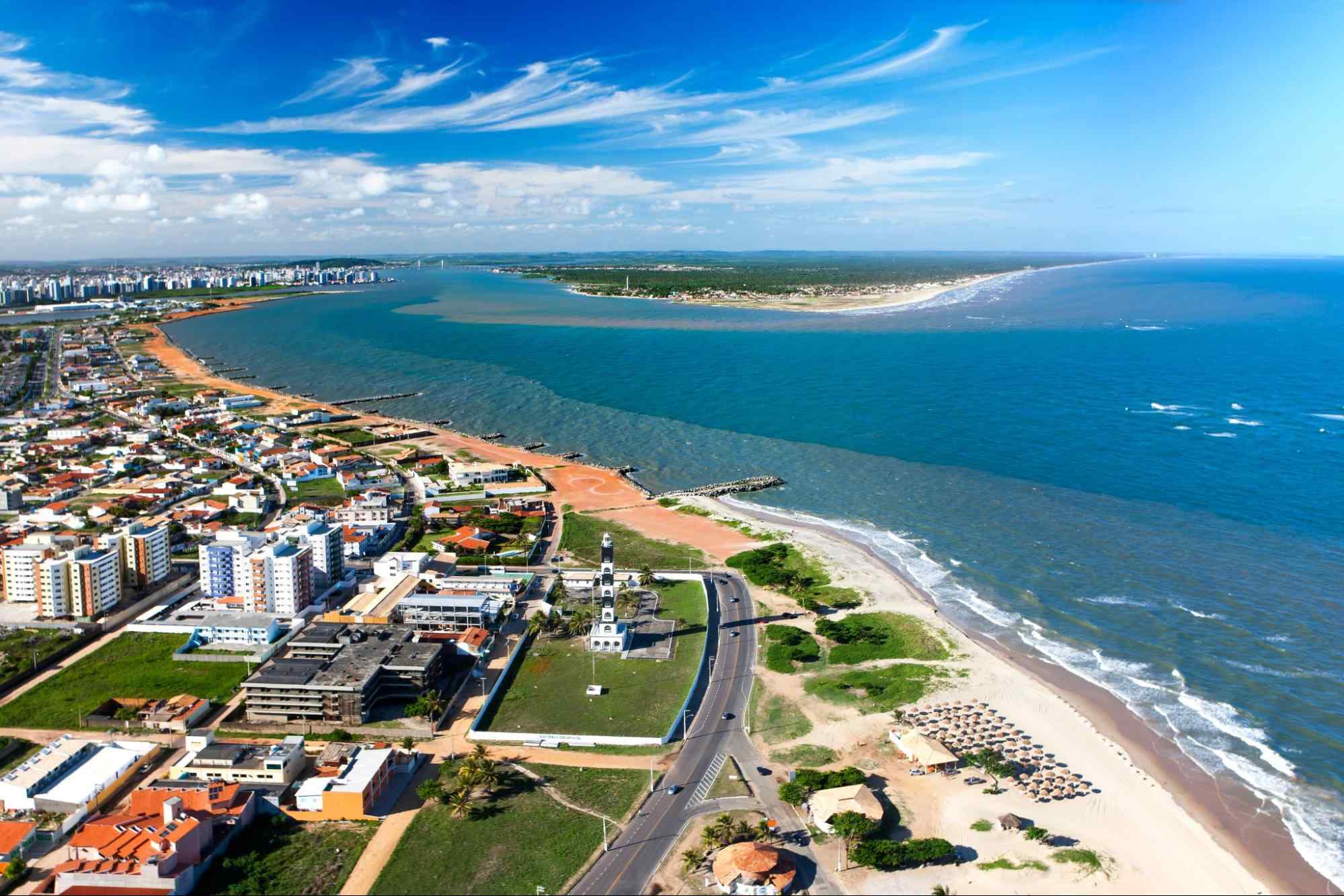 Vista aérea da zona costeira de Aracaju, capital de Sergipe. A paisagem urbana é composta por prédios baixos e áreas verdes que encontram uma longa faixa de areia dourada. Depois dela, aparece um mar azul que se estende até o horizonte.