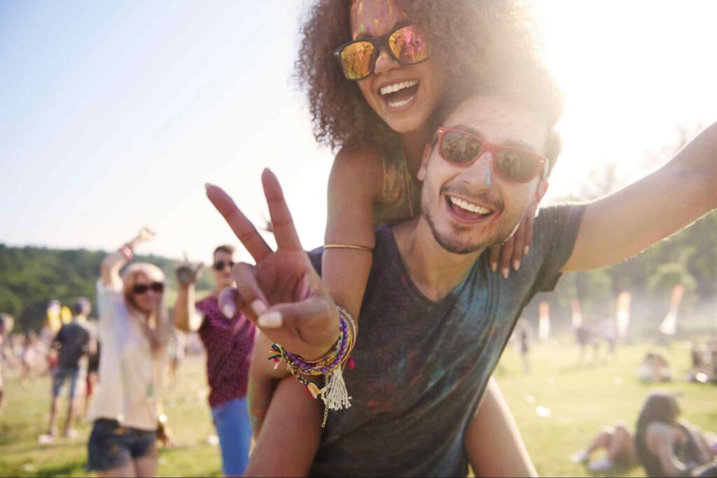 Foto de pessoas se divertindo em festival ao ar livre, com uma mulher nas costas de um homem em destaque.
