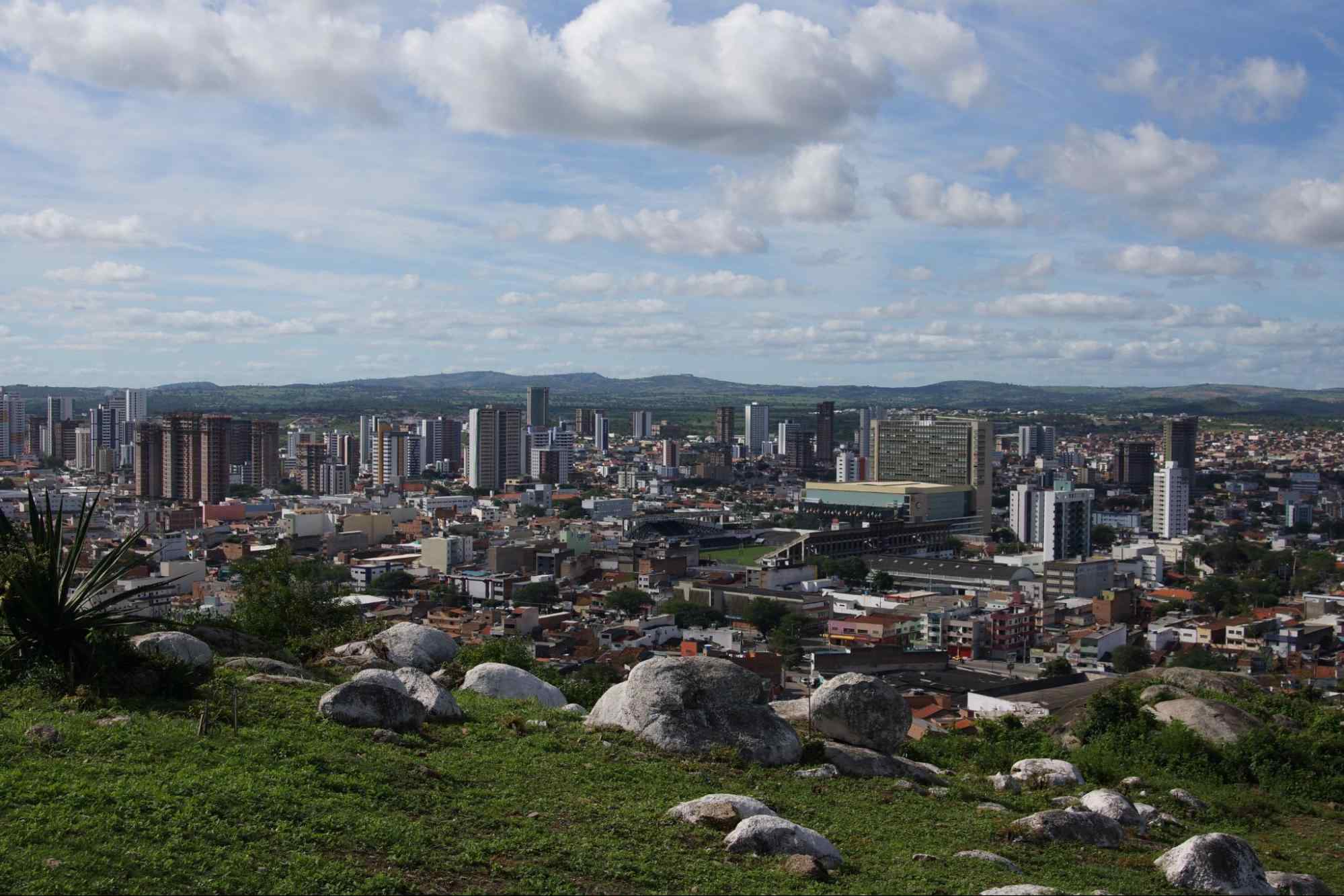 Vista aérea de Caruru, PE. A paisagem urbana é formada majoritariamente por construções baixas, com alguns prédios maiores, e rodeada por serras verdes.