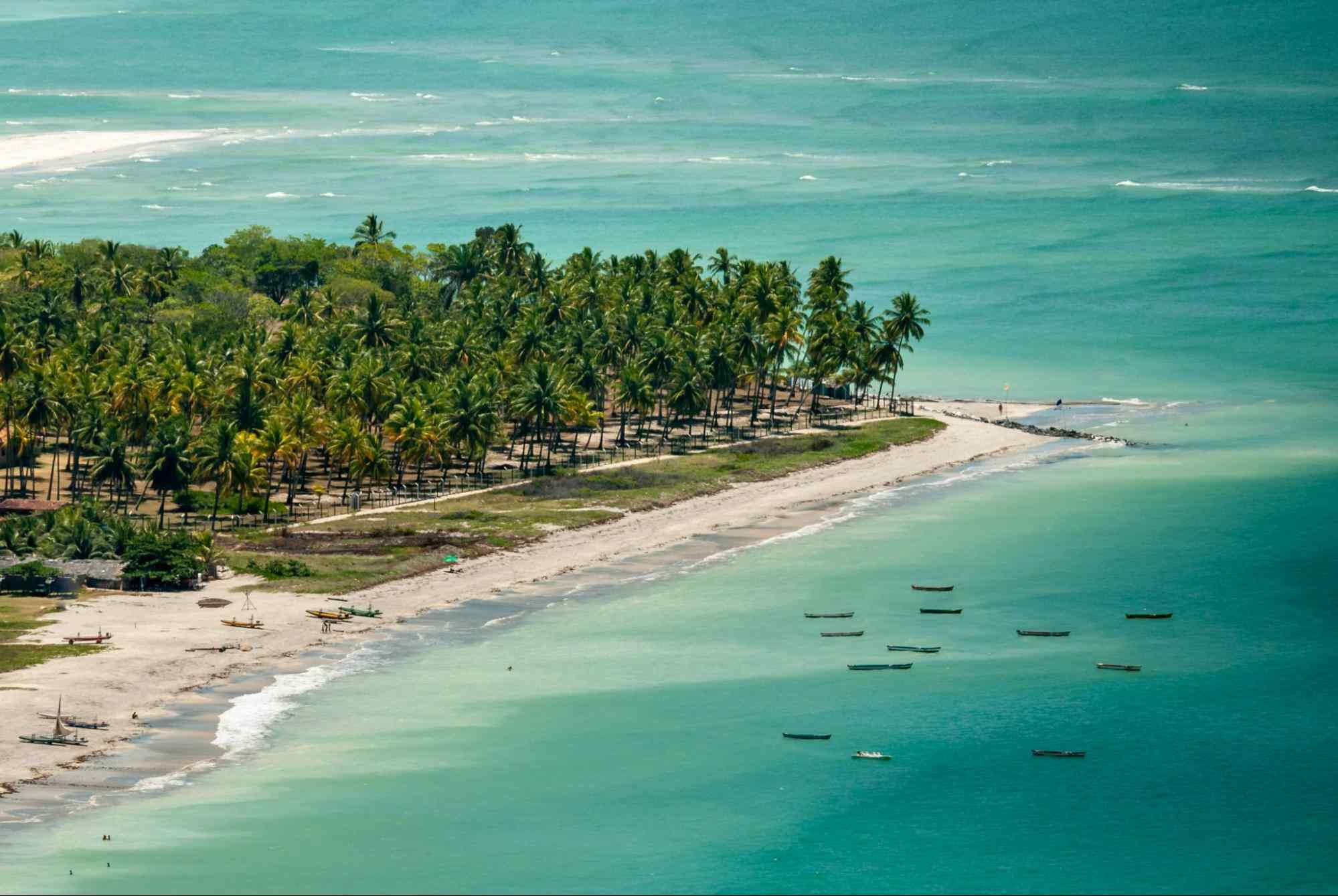 Foto aérea de parte da Ilha de Itamaracá. Pontal banhado pelo mar azul claro, com faixa de areia branca, gramado e diversos coqueiros.