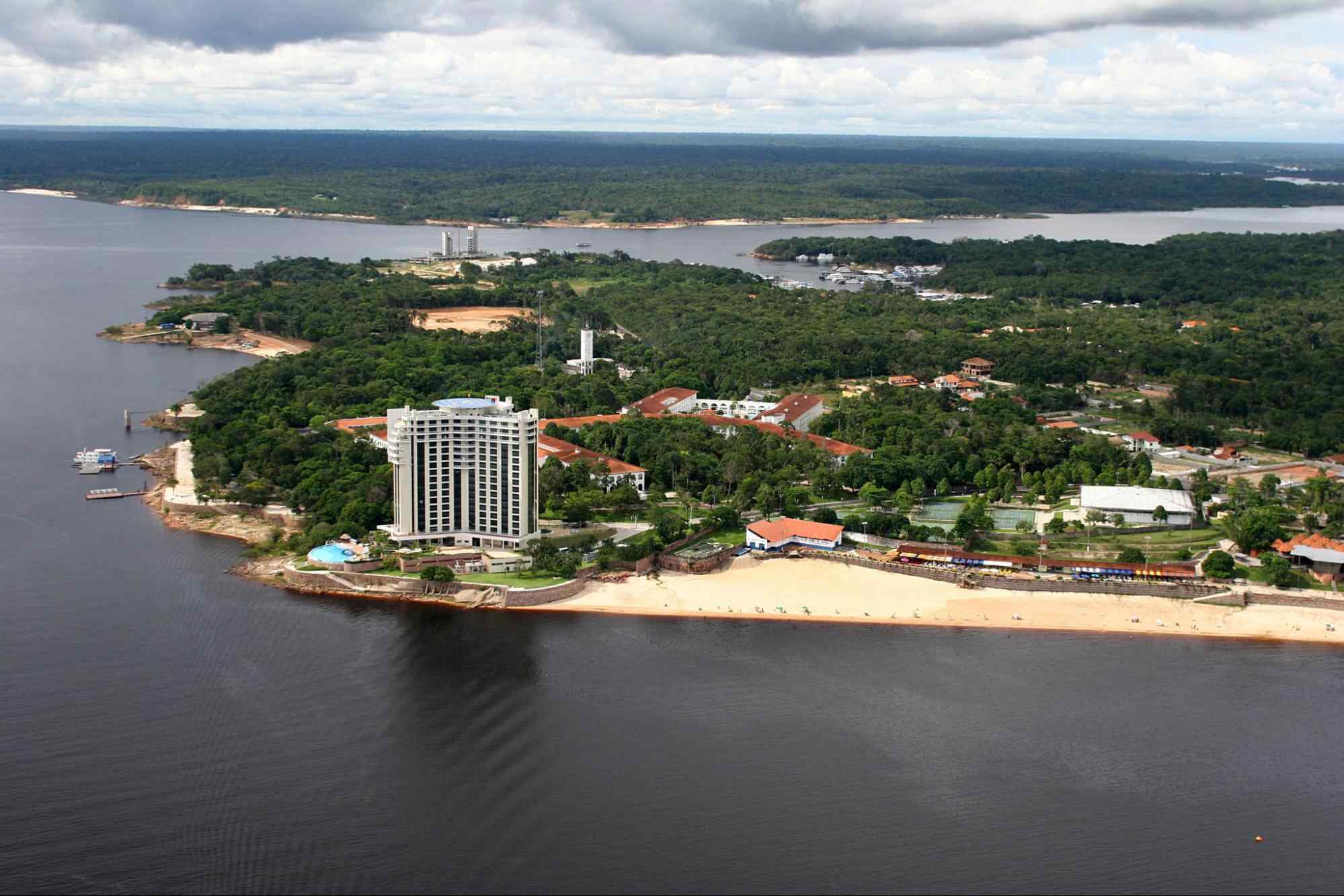 Vista aérea de uma parte de Manaus. Os trechos de terra se intercalam com as águas escuras do rio e são densamente arborizados, com algumas construções baixas e apenas um prédio moderno e grande.