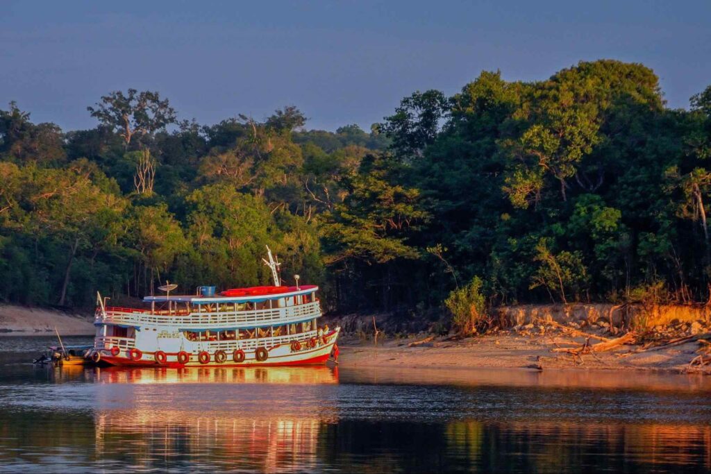 Embarcação de dois andares flutuando no rio em Manaus. Ela se aproxima da terra, onde há um trecho de mata verde e densa.