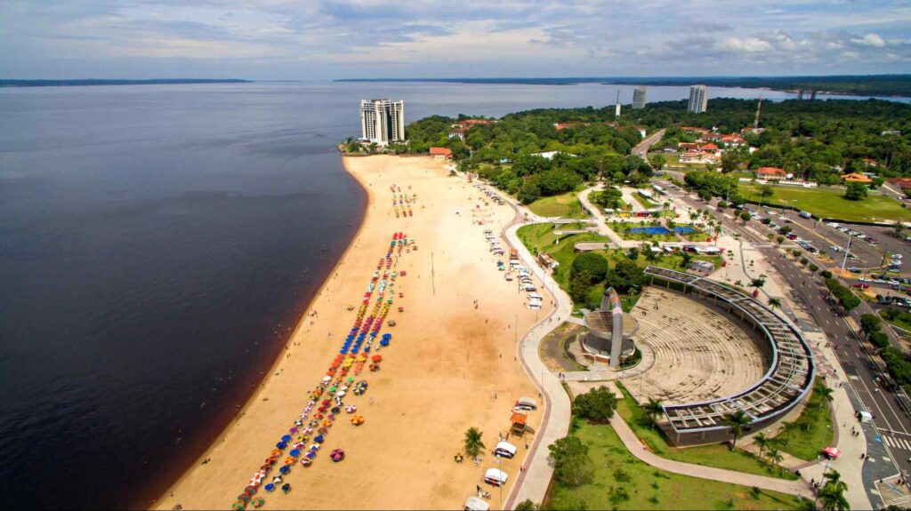 Vista aérea da Praia de Ponta Negra, Manaus. A larga faixa de areia dourada é limitada pelas águas negras do rio e por um longo calçadão onde fica um anfiteatro. Há alguns guarda-sóis e barraquinhas espalhados pela areia.

