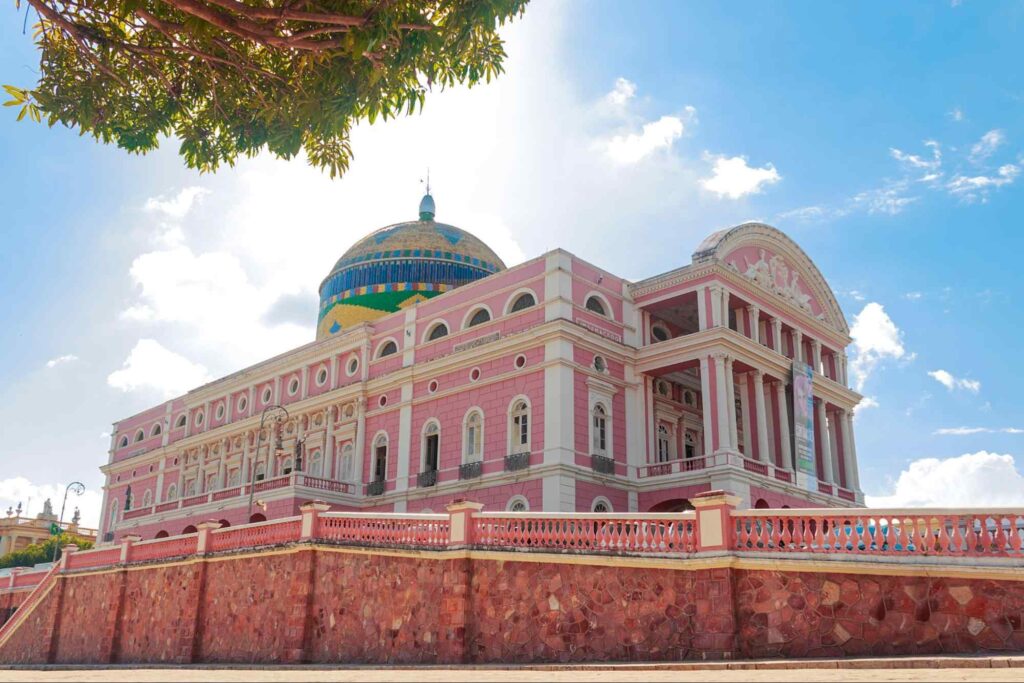 Teatro Amazonas, em Manaus. A grande construção de arquitetura renascentista tem uma fachada de cor rosa marcada por grandes colunas e muitas janelas. No topo, há uma cúpula com mosaicos em azul, verde e amarelo.
