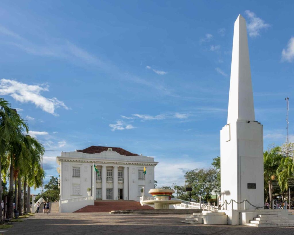 Palácio Rio Branco, uma antiga mansão de fachada branca com janelas quadradas e grandes colunas. À frente da construção, está o monumento monolítico conhecido como Obelisco do Acre
