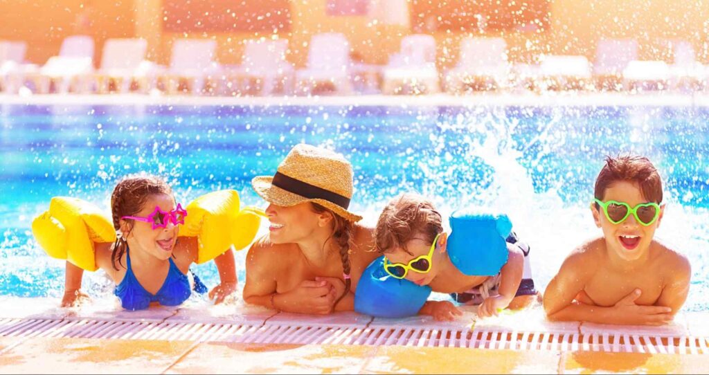Uma mulher e três crianças apoiados na borda de uma piscina, com os corpos parcialmente submersos. As crianças usam óculos escuros de armações coloridas e têm boias nos braços