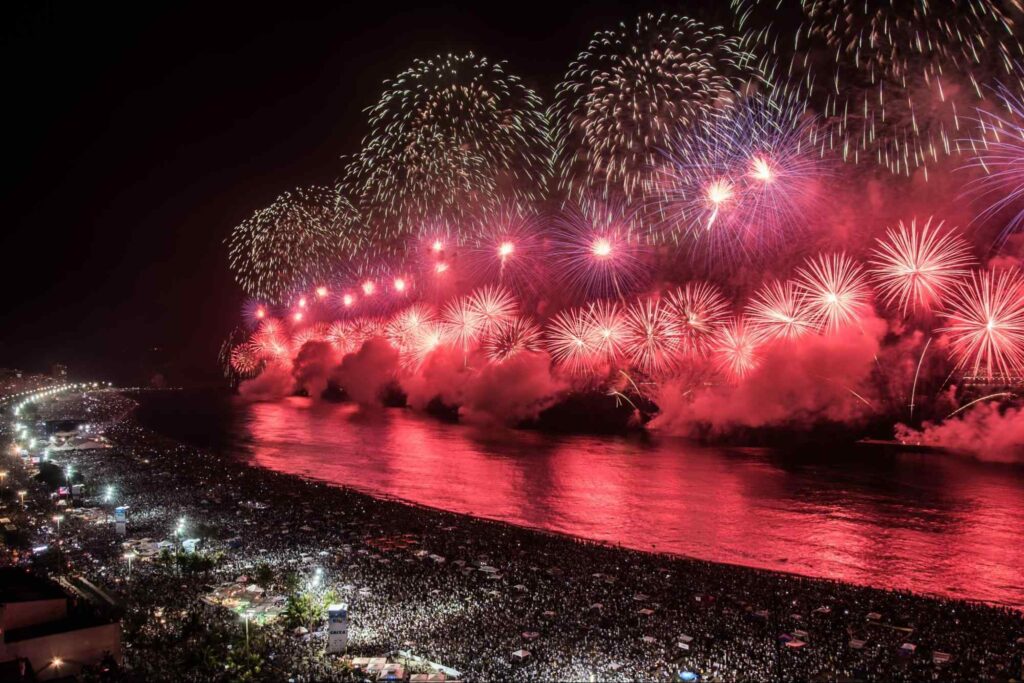 Fogos de artifício em praia brasileira, durante a virada do ano. Fogos vermelhos, refletindo no mar, com inúmeras pessoas na areia, um palco iluminado e luzes azuis projetadas no céu