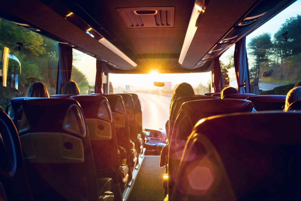 Vista da parte interior traseira de um ônibus, com alguns bancos e partes das cabeças dos passageiros, com o sol se pondo na frente do ônibus