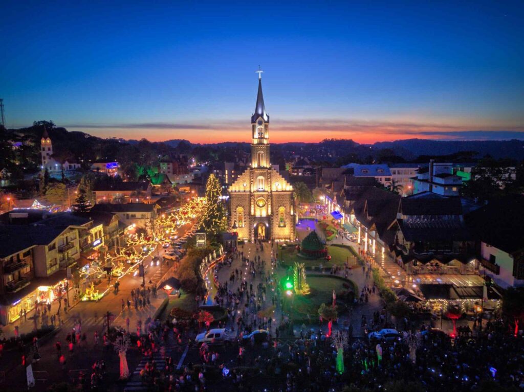 Vista aérea de Gramado, RS, à noite. As casas de estilo arquitetônico alemão estão decoradas por luzes de Natal, assim como a Paróquia São Pedro, um grande templo de pedra. Um grande número de pessoas circula pela praça da igreja