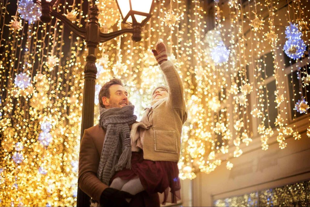 Homem jovem segurando uma criança, que parece ser sua filha, sob um arranjo de luzes pendentes de Natal