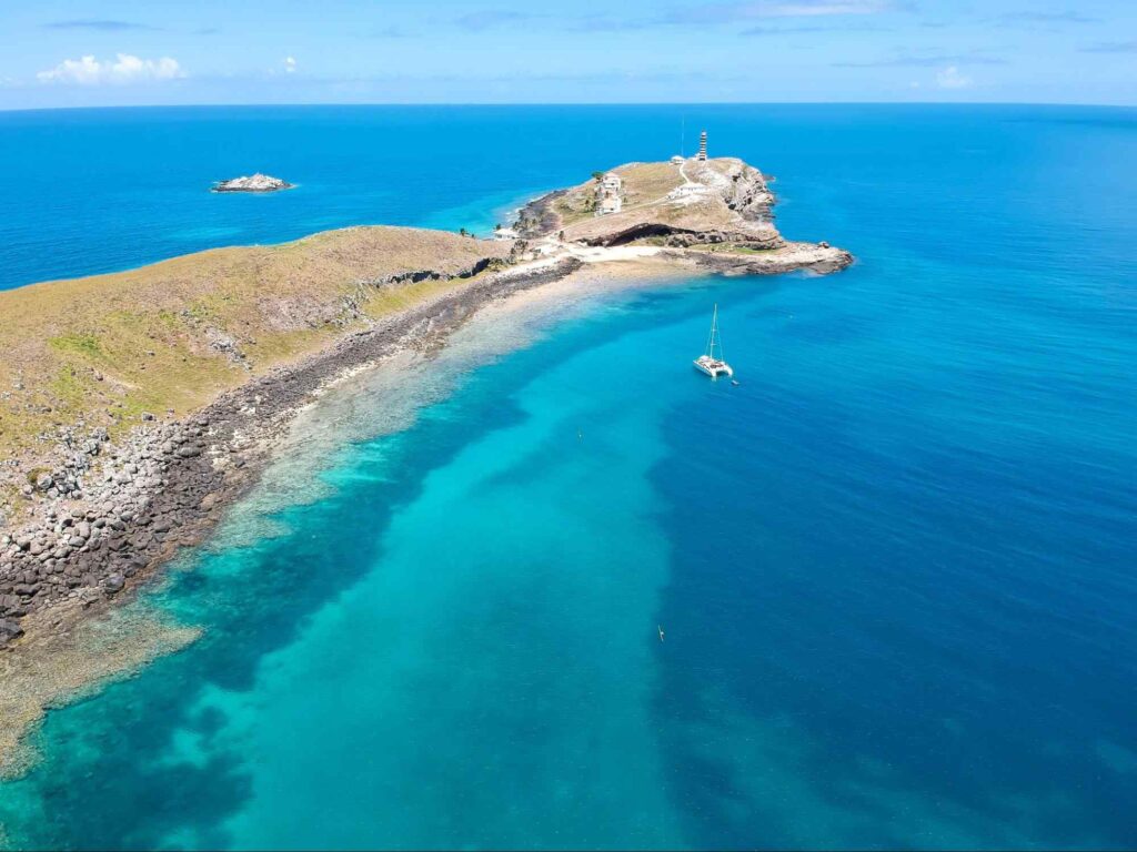 Foto aérea de um pontal rochoso em Abrolhos, Bahia. O estreito trecho de pedra é rodeado pelas águas de azul intenso do mar, em que uma pequena embarcação branca navega