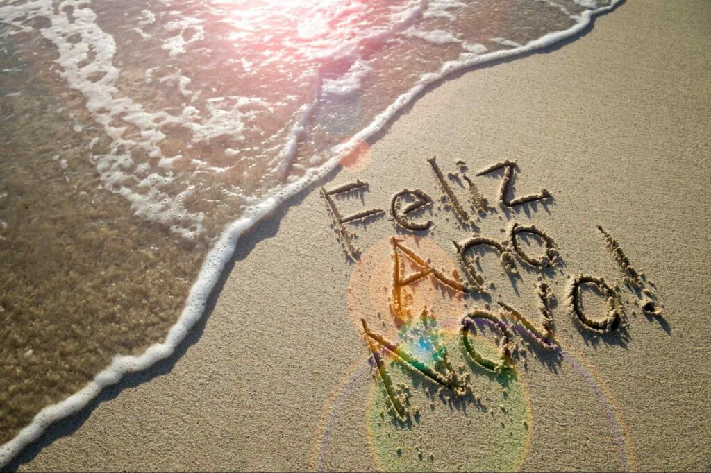 Foto de escrita na areia da praia, com água do mar quase encostando na frase “Feliz ano novo!