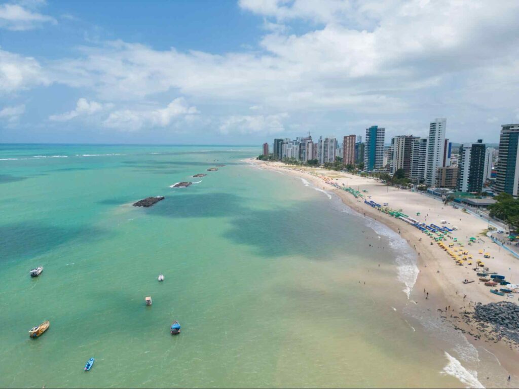 Imagem aérea da Praia de Candeias, Jaboatão dos Guararapes PE. Mar azul ao lado esquerdo da imagem, com extensa faixa de areia repleta de guarda-sóis coloridos e prédios de diferentes tamanhos na orla