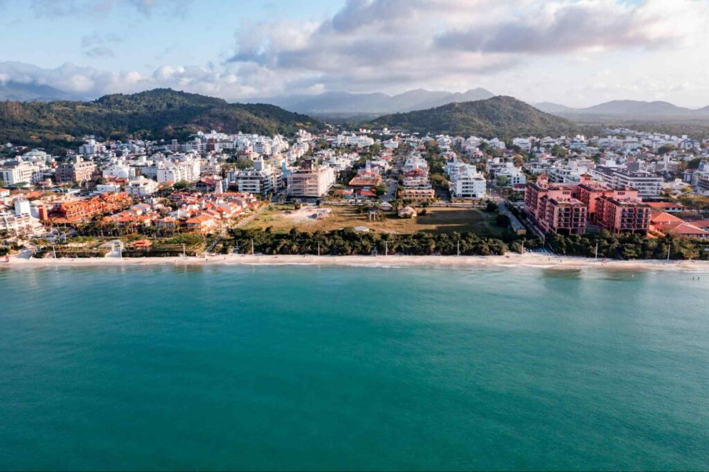 Imagem panorâmica da Praia de Jurerê Internacional, com o ponto de vista do mar para a cidade. Mar azul, em frente a extensa faixa de areia, com árvores,prédios e casas atrás. No horizonte, montanhas verdes