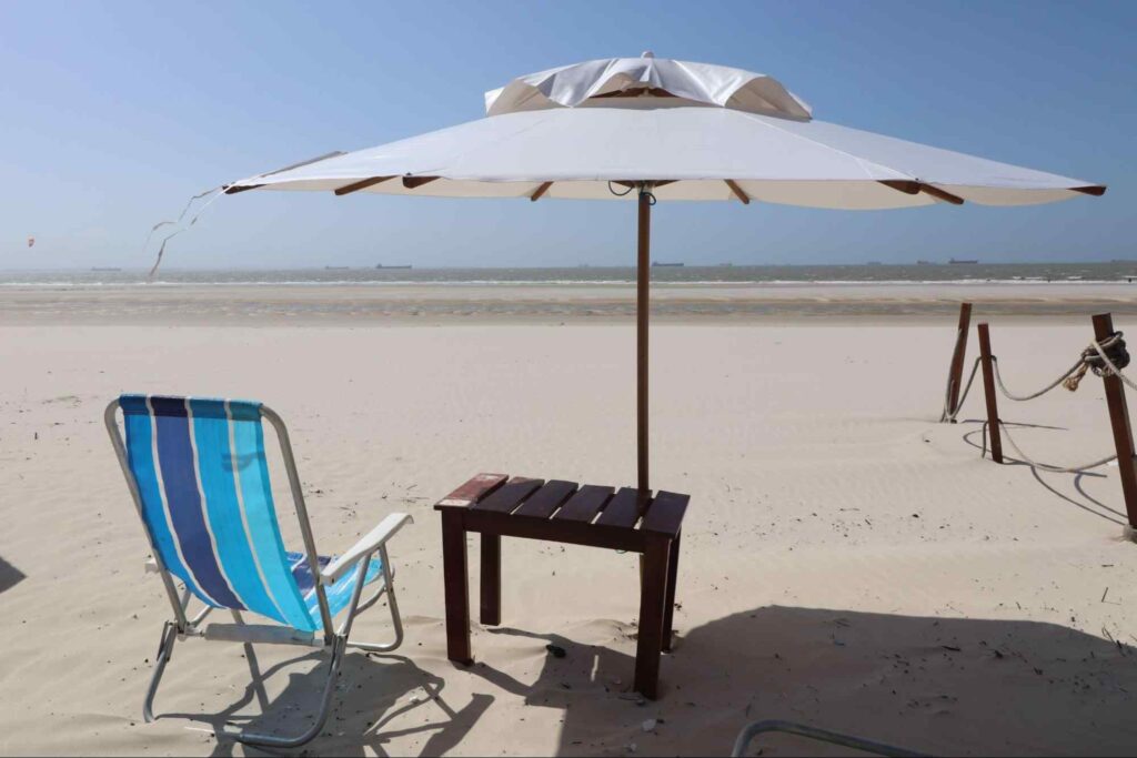 Cadeira de praia ao lado de uma mesinha de madeira, sob um guarda-sol, na Praia de Araçagi. A areia branca e limpa é limitada pelo mar e está completamente vazia