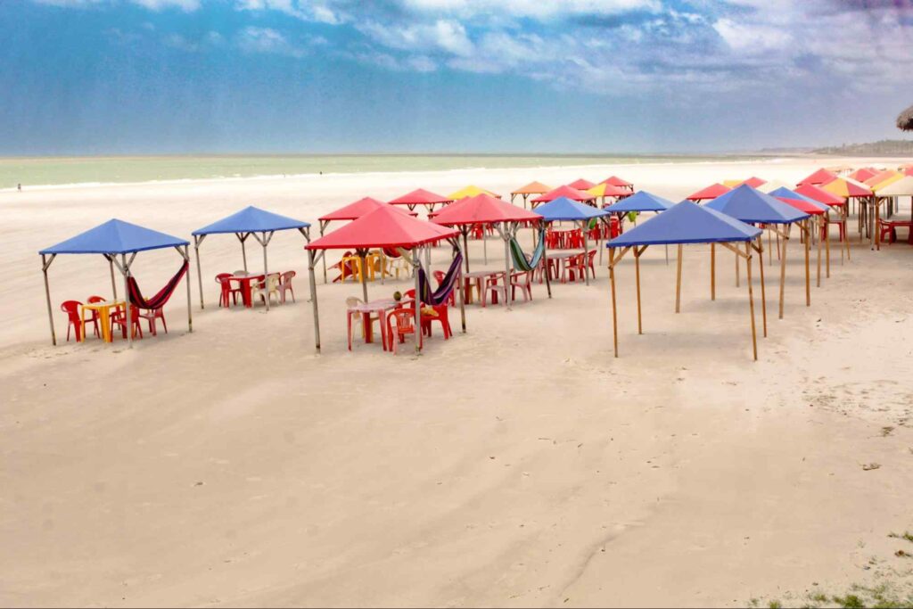 Barraquinhas com toldos coloridos cobrindo mesas, cadeiras e redes na areia de uma das praias de São Luís. Ao fundo, aparece o mar esverdeado, que se estende até o horizonte