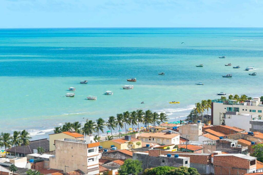 Fotografia aérea de Maragogi, AL. A cidade tem construções baixas, como casas e bangalôs de frente para o mar. Pequenas embarcações flutuam no azul da água, que se estende até o horizonte.