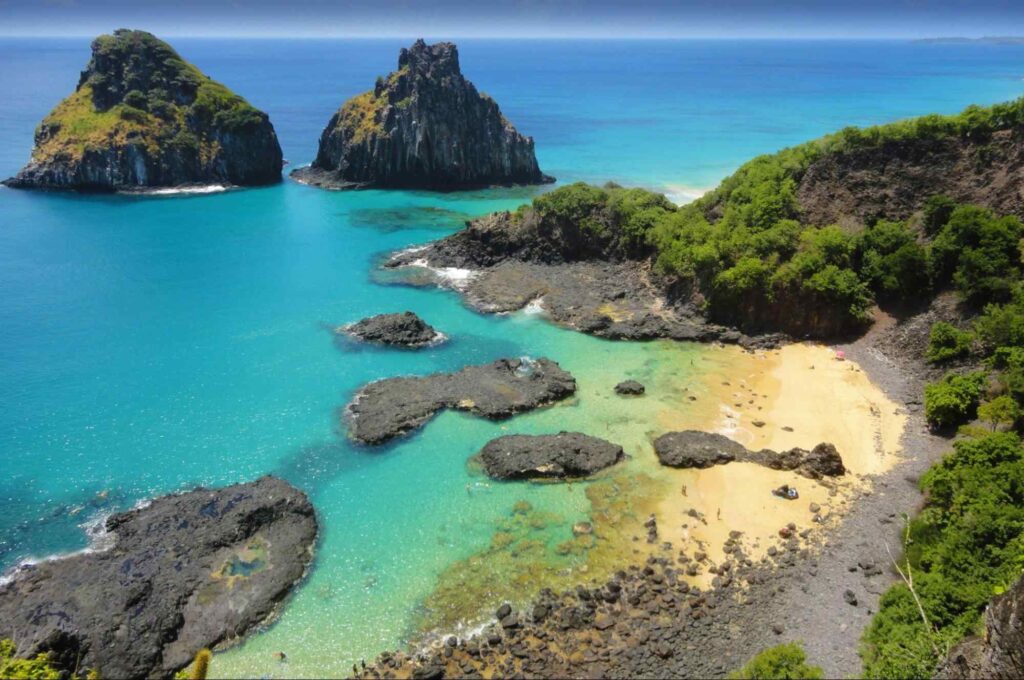 Foto de parte da praia de Fernando de Noronha, PE. Formações rochosas em meio a água azul cristalina do mar, com pequena faixa de areia ao lado direito da foto, cercada por mais formações rochosas