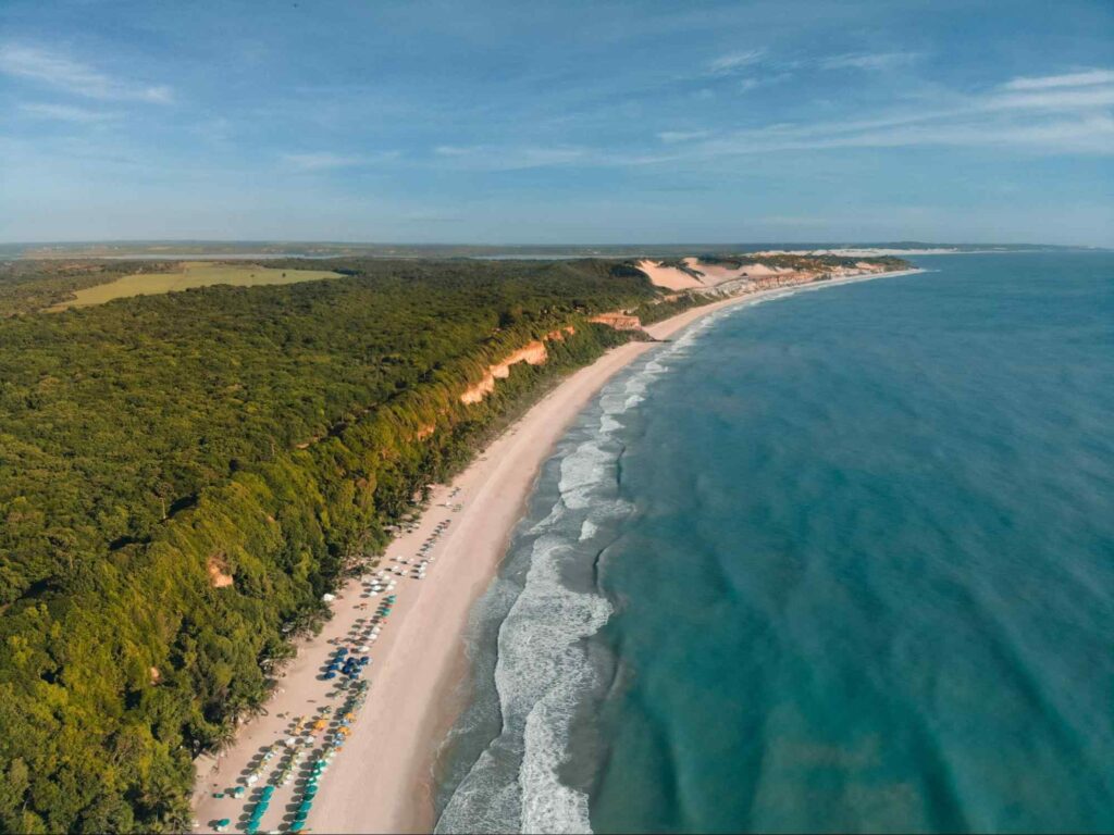 Foto panorâmica da Praia da Pipa, RN. Grande área verde ao lado esquerdo da foto, em frente à extensa faixa de areia que fica em frente ao mar azul.
