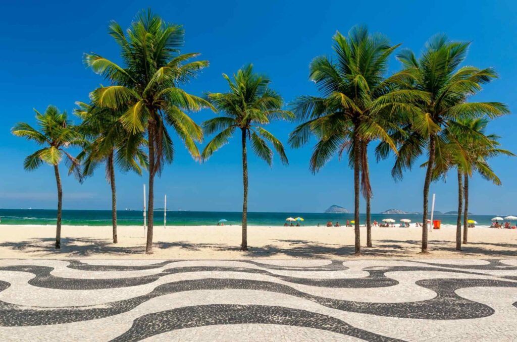 Foto da Praia de Copacabana, mostrando o mosaico da calçada em frente à palmeiras, faixa de areia e o mar azul no horizonte