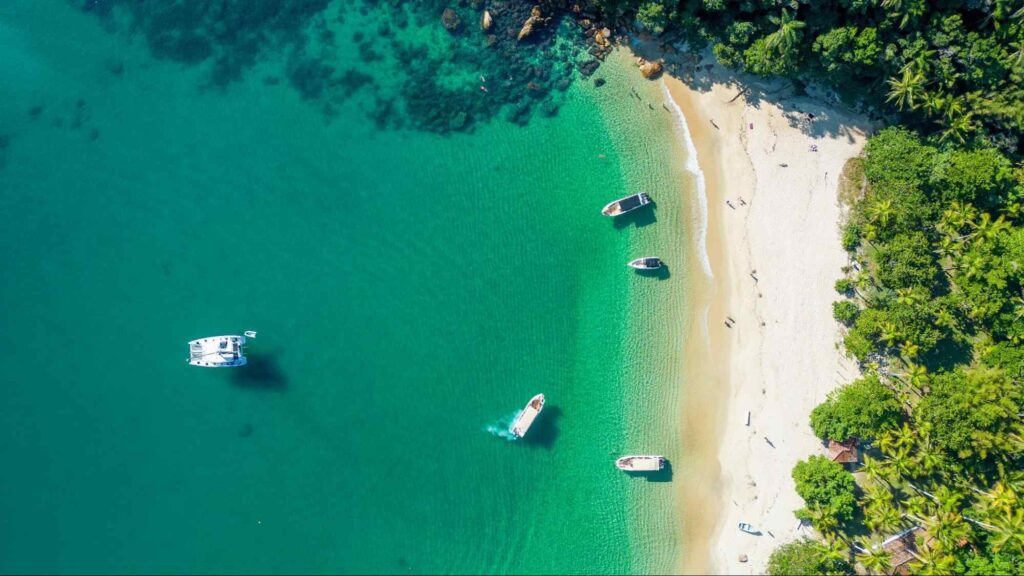 Praia de Angra dos Reis vista de cima. Mar cristalino esverdeado com alguns barcos, em frente a pequena faixa de areia, cercada pela vegetação natural