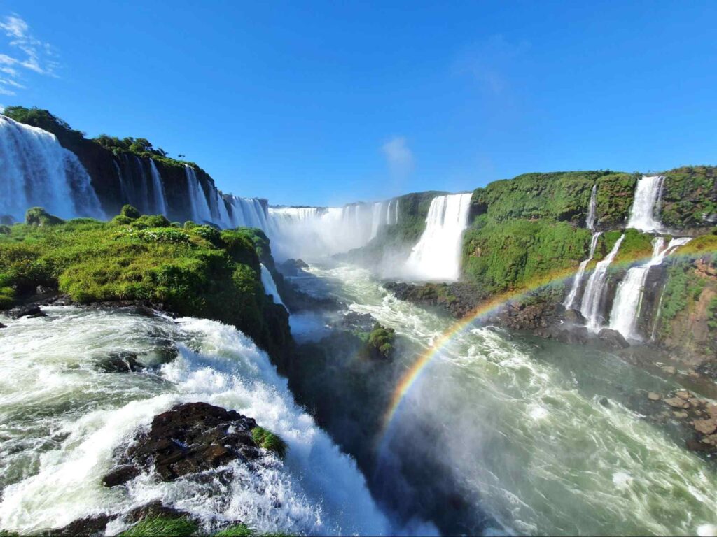 Cataratas do Iguaçu em dia de céu azul e limpo, com as quedas formando um arco-iris no canto direito da foto