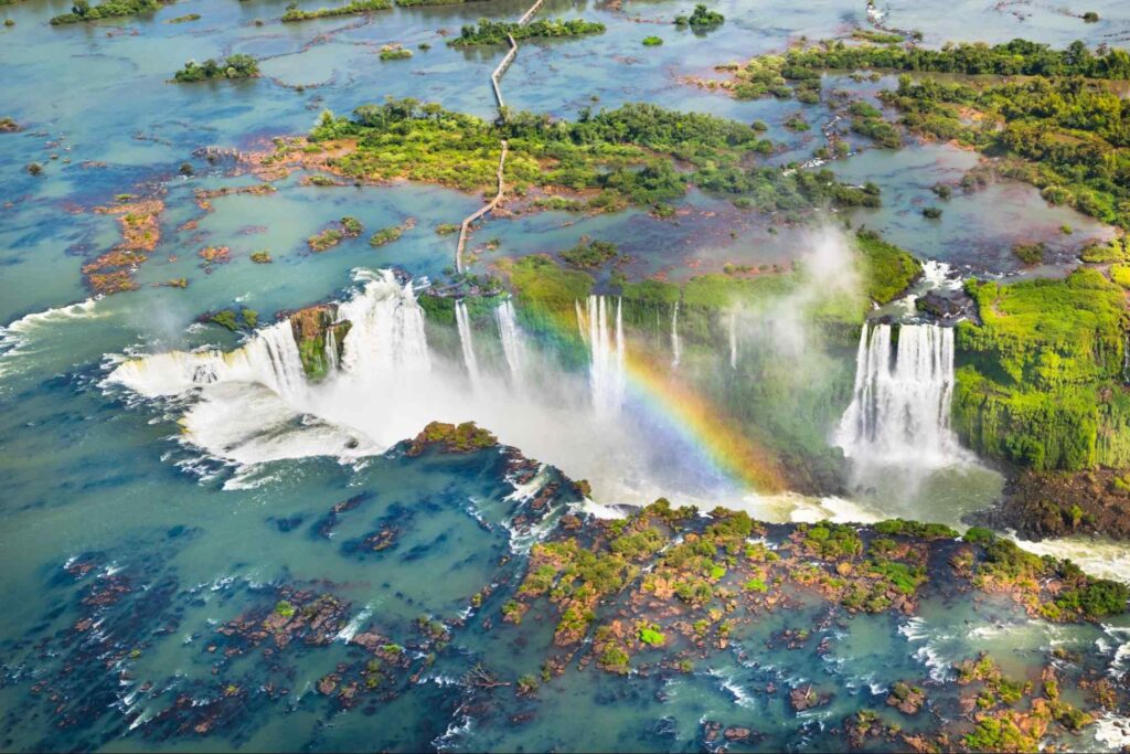 Vista aérea das Cataratas do Iguaçu, no Parque Nacional do Iguaçu. A água azul se acumula em um planalto e divide espaço com alguns trechos de vegetação verde. Ela despenca por uma falha geológica em formato de U, formando um grande complexo de cachoeiras.