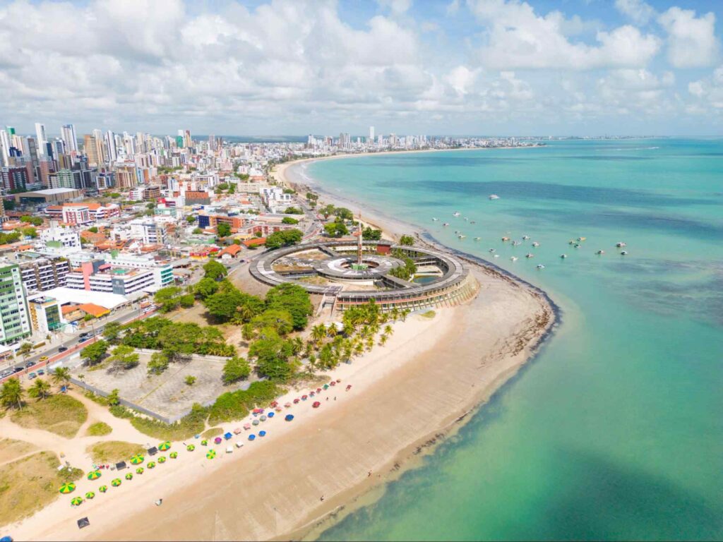 Vista aérea do litoral de João Pessoa, PB. A paisagem urbana é composta por muitos prédios modernos de tamanhos variados e delineada por uma longa faixa de areia que a separa do mar azul