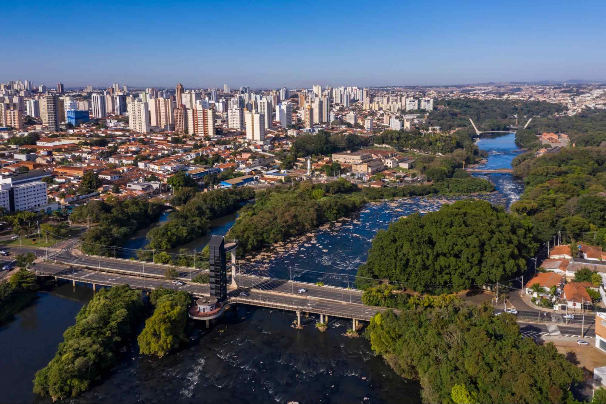 Vista aérea de Piracicaba, SP. A cidade é cortada por áreas densamente arborizadas e pelas águas do Rio Piracicaba. A paisagem urbana é composta tanto por construções baixas quanto por prédios altos e modernos