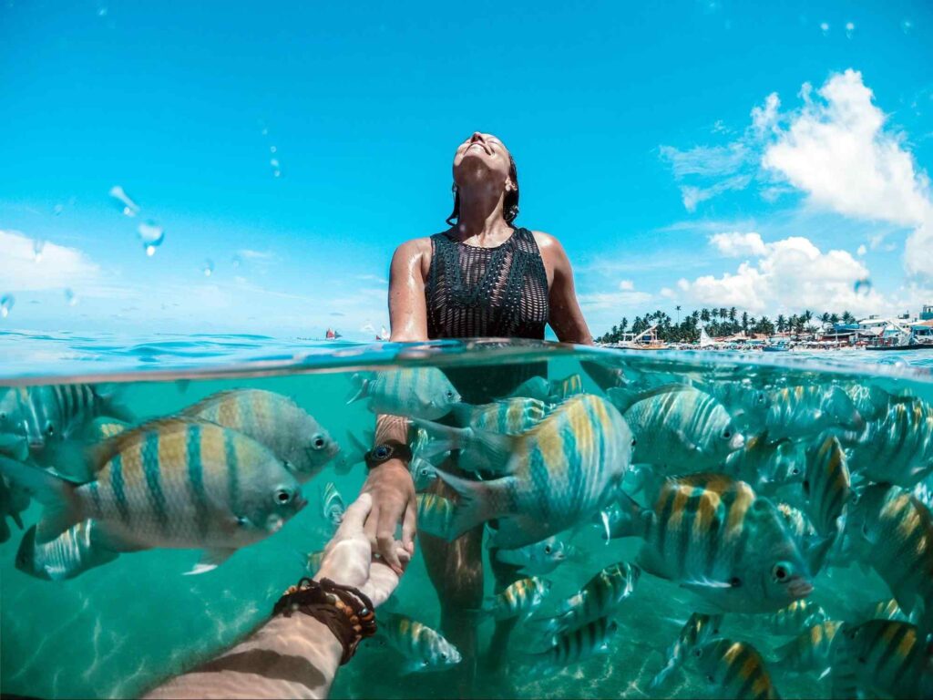Banhista mergulhando nas águas cristalinas do mar. Ela está submersa até a altura do tórax e rodeada por pequenos peixes azulados