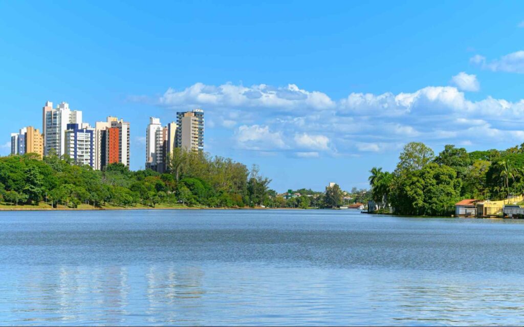 Paisagem da cidade de Londrina, com Lago Igapó em destaque na foto. Prédios de diferentes tamanhos ao lado esquerdo da foto, com árvores em frente aos prédios, na margem do lago