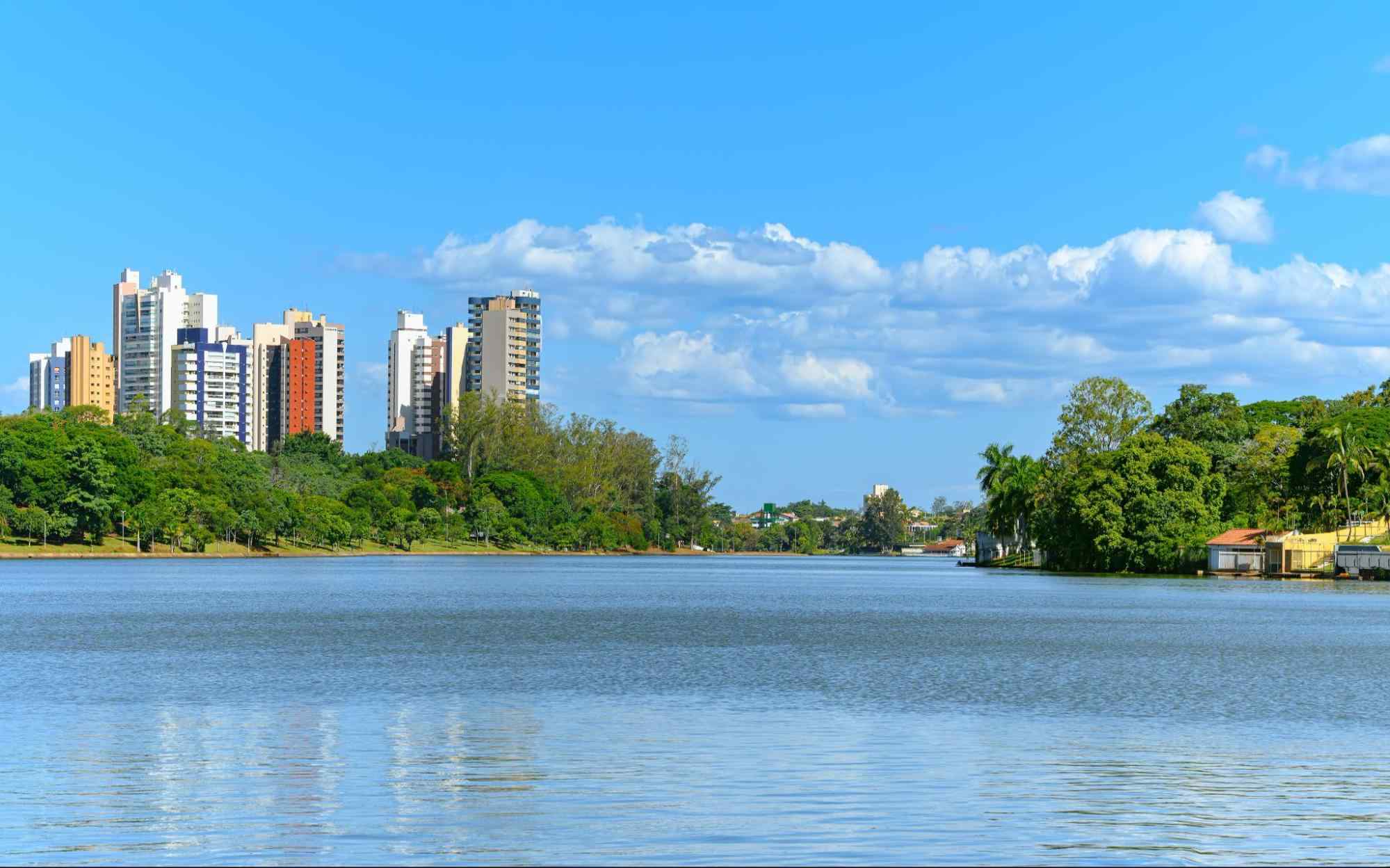 Paisagem da cidade de Londrina, com Lago Igapó em destaque na foto. Prédios de diferentes tamanhos ao lado esquerdo da foto, com árvores em frente aos prédios, na margem do lago