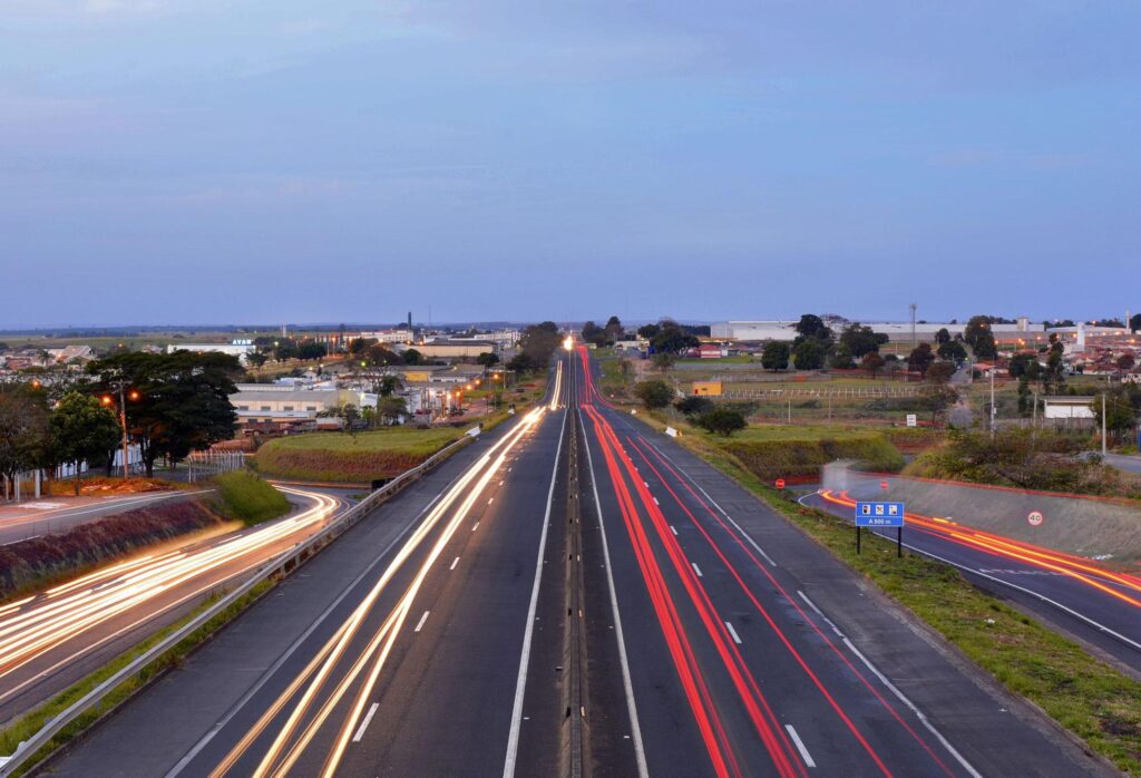 Rodovias que dão acesso a Marília, SP. As duas longas estradas são cortadas por feixes de luz coloridos, deixados pelos faróis dos automóveis que circulam em alta velocidade. Ao fundo, aparece a paisagem urbana, composta principalmente por galpões e outras construções baixas