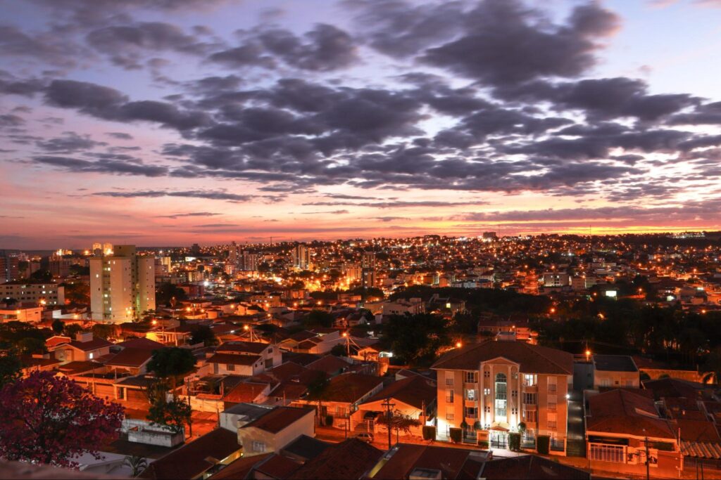 Vista aérea panorâmica da cidade de São Carlos SP ao entardecer. Luzes amarelas dos postes das ruas em contraste ao céu azul passando para o alaranjado pôr do sol.