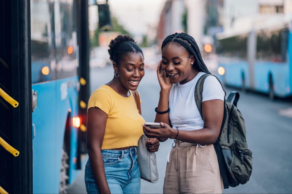Duas jovens sorriem e olham para a tela do celular que uma delas segura. Ao fundo e na lateral, aparecem dois ônibus de viagem azuis.