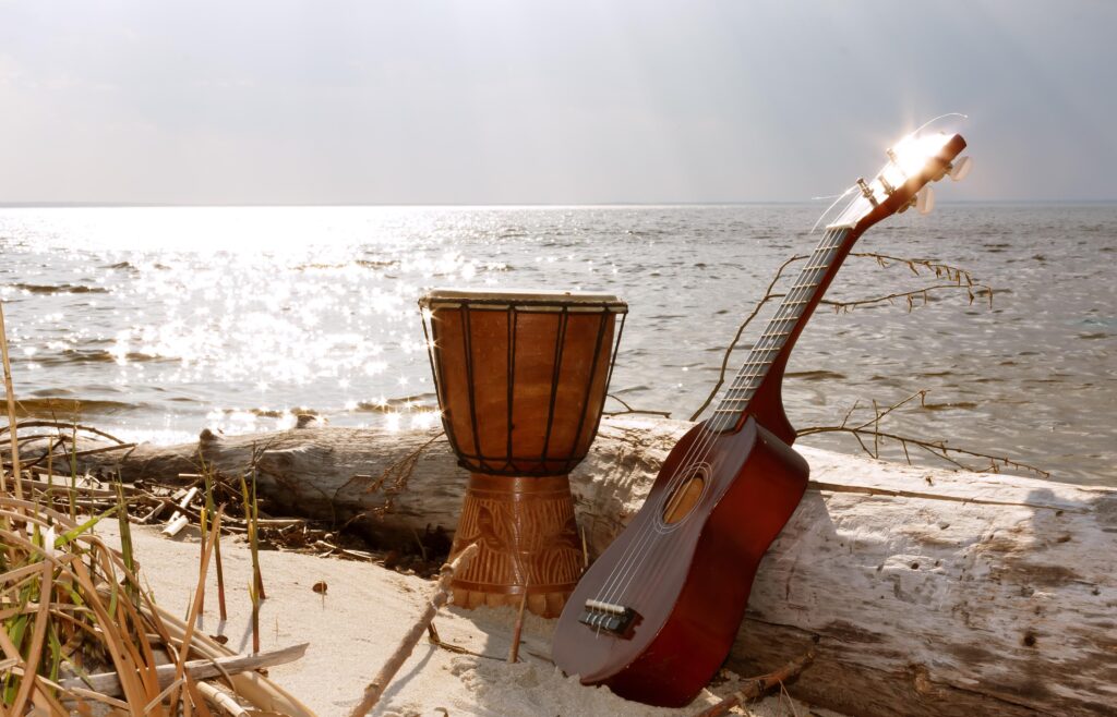 Batuque tradicional e um violão encostado em um tronco na areia com o rio ao fundo
