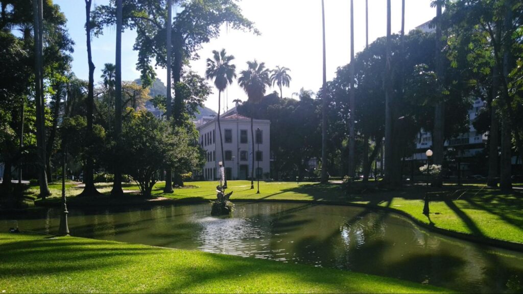 Lago no jardim do Palácio do Catete. As águas escuras estão rodeadas por um gramado verde e por árvores altas, como palmeiras. O dia está ensolarado. Ao fundo, aparece uma parte da fachada do palácio.