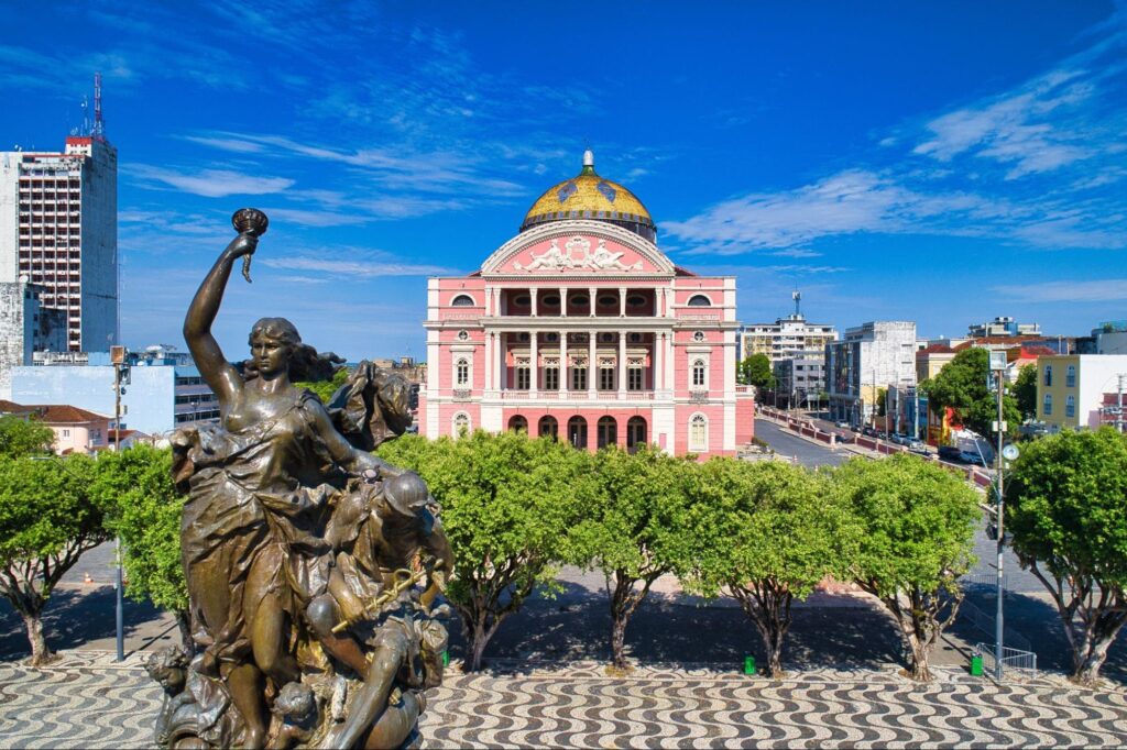 Teatro Amazonas, em Manaus, visto de cima. A grande casa de espetáculos de três andares tem uma arquitetura em estilo renascentista, com colunas e arcos em toda a fachada cor-de-rosa. Em frente ao teatro, aparece uma praça arborizada com destaque para uma estátua de cobre