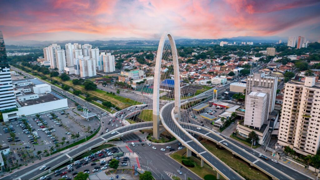 Vista aérea da principal rodovia com ponte estaiada da cidade de São José dos Campos ao entardecer. Ao horizonte, céu azul coberto por nuvens em tons rosados e alaranjados.