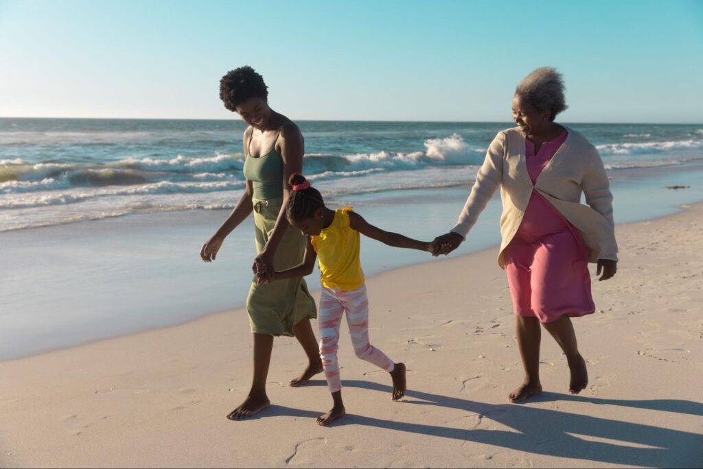 Família composta por filha, mãe e avó. As três caminham de mãos dadas na areia da praia, em frente ao mar.